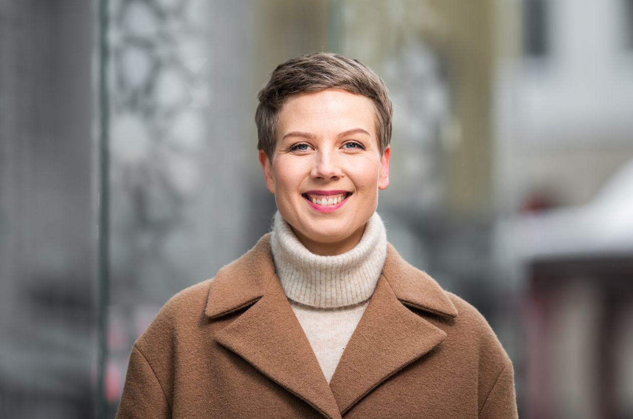 Vihreiden kansanedustaja Jenni Pitko katsoo suoraan kameraan taustallaan rauhallinen kaupunkimiljöö.