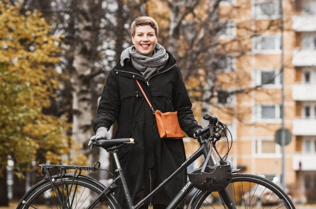 Jenni Pitko seisoo pyörän kanssa keltaisen kerrostalon edustalla syyspuiden alla.