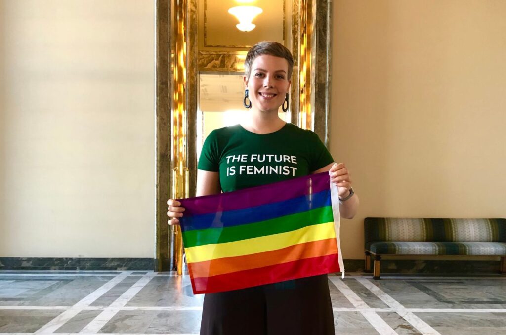 Vihreiden kansanedustaja Jenni Pitko katsoo suoraan kameraan eduskuntatalolla ja hymyilee kädessään pride-lippu.